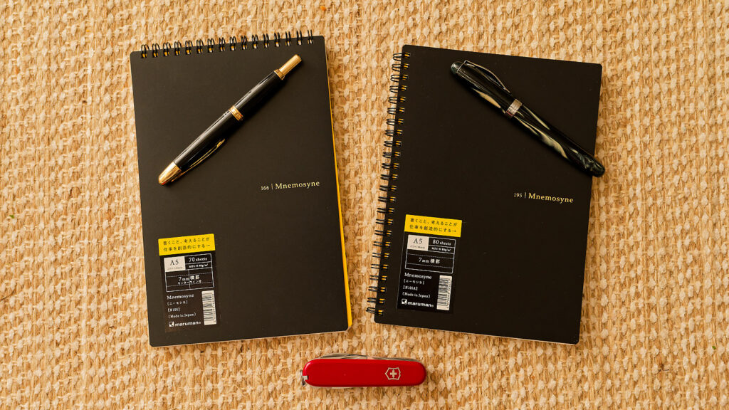 Two maruman Mnemosyne notebooks next to each other on a woven table runner. They each have a fountain pen on them. At the bottom of the photo is a swiss army knife. 