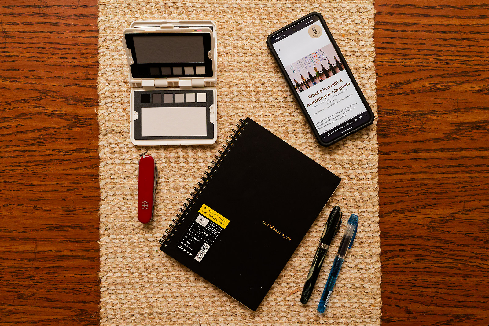 A Maruman Mnemosyne notebook with a cell phone, a swiss army knife, two fountain pens, and a color checker next to it on a table