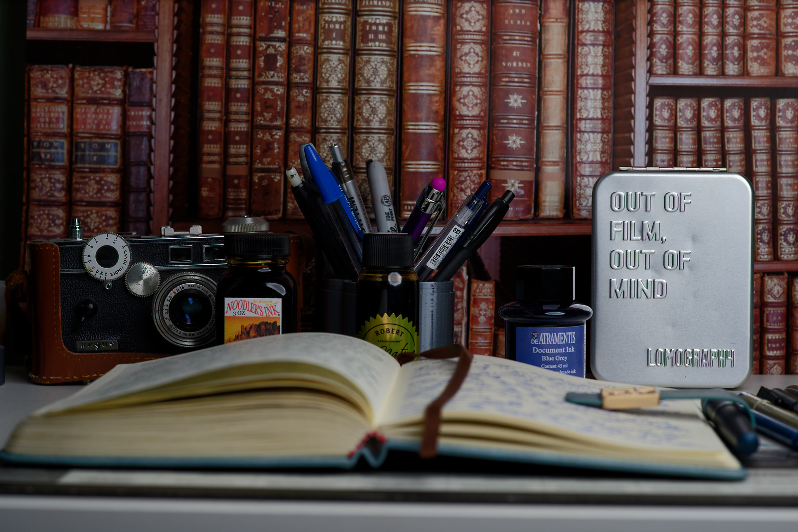 A journal on a desk filled with pens, analog photo equipment, and set against a book case background