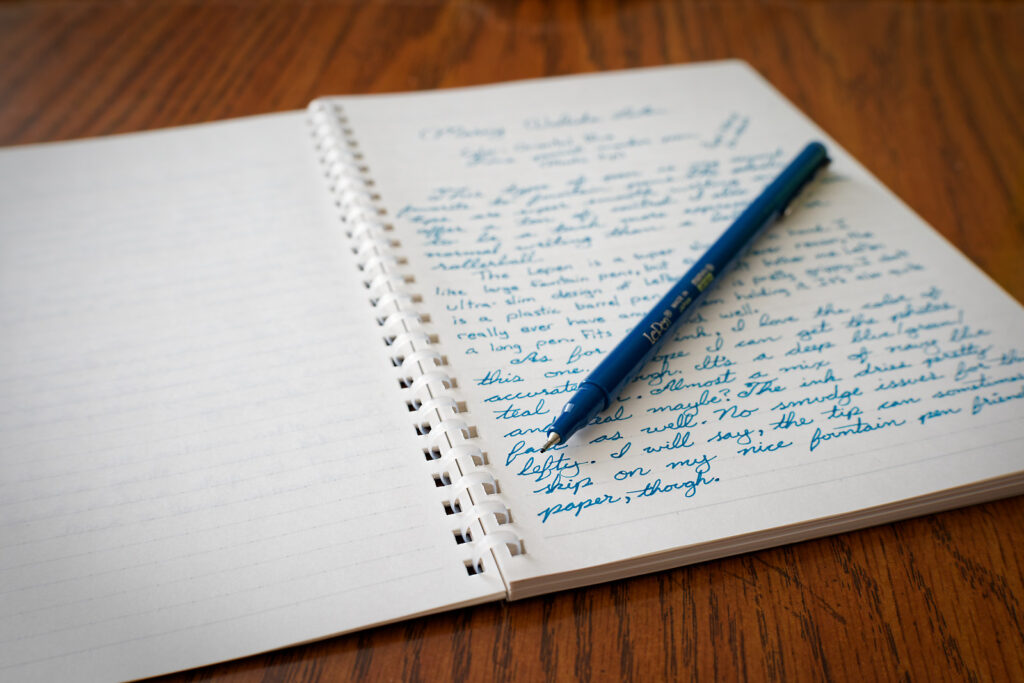 A photo of the tip-end of a Marvy Le Pen placed onto a Kokuyo Biz notebook with a writing sample. 