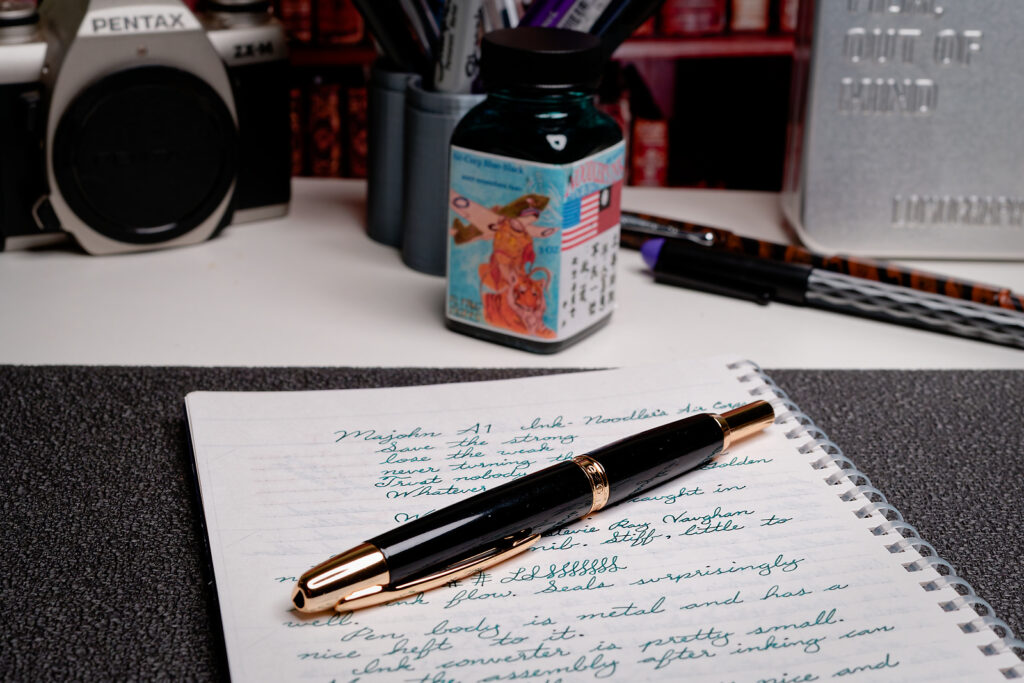 A photo of a Majohn A1 black and gold fountain pen sitting on a notebook with writing samples under it. 