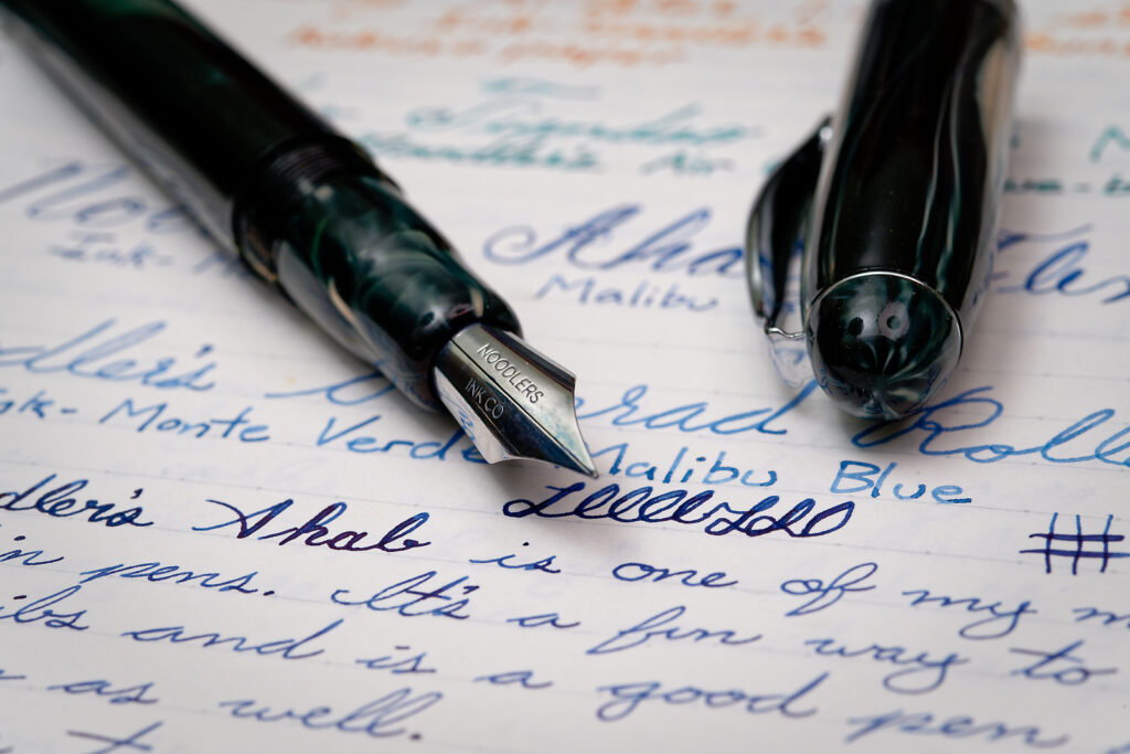A photo of a Noodler's Ahab flex fountain pen with the nib up close to the camera and the cap beside it. It shows a writing sample on paper under the pen. 