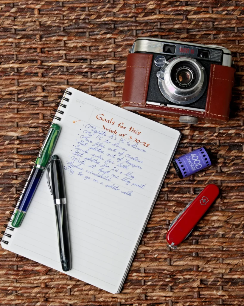 A flat lay photography of a Noodler's Konrad and a Noodler's Ahab fountain pens laying on a Maruman Mnemosyne with a To-do list written by those pens. Next to that is a Swiss Army knife, a roll of Cinestill 35mm film, and an old Kodak camera.  
