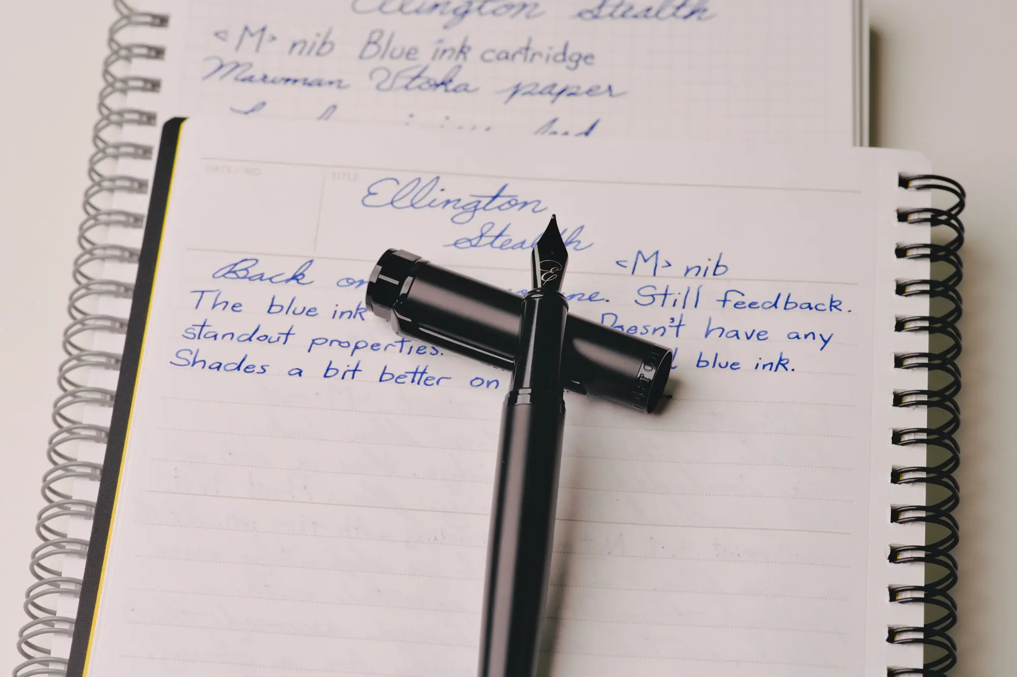 A matte black Ellington Stealth fountain pen with it's section resting on top of the cap. The whole thing is placed on top of a couple of notebooks featuring writing done with the pen.