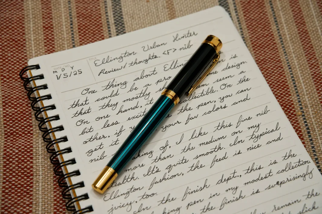 This photo shows an Ellington Pens fountain pen sitting on a lined piece of paper. The pen is the Urban Hunter model, featuring a gold colored grip section, clip, nib, can cap bands. The body is a deep teal/green color with black, vertical stripes.